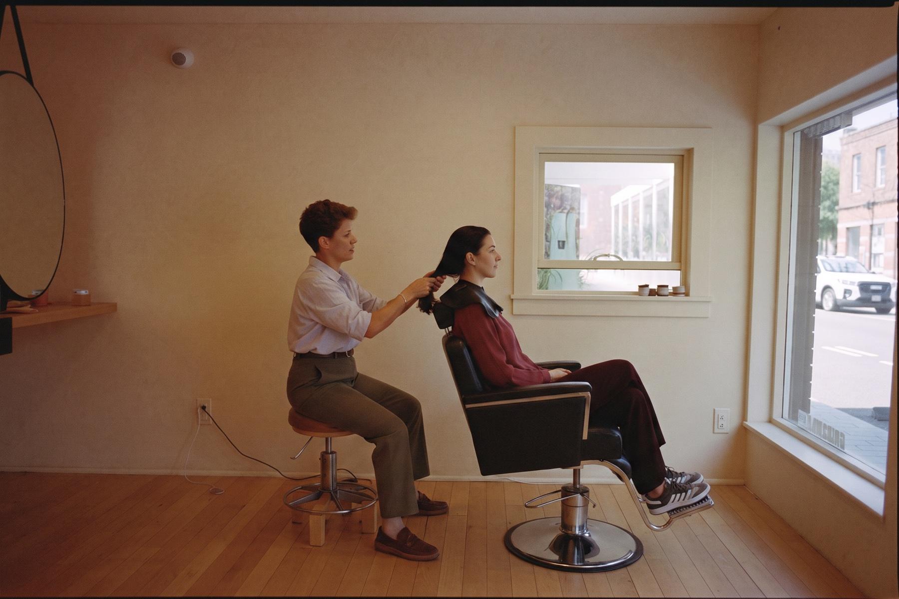 A stylist seated on a low stool consulting with a guest seated separately in the salon chair, at eye level