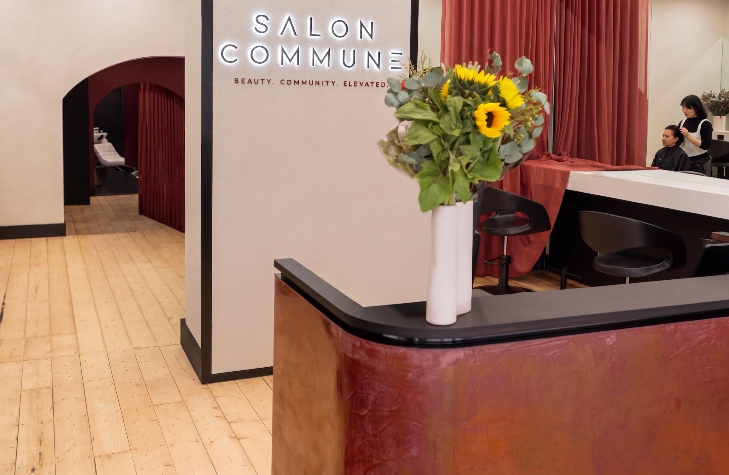 The curved oxblood reception counter with a sunflower and native arrangement, beside the backlit Salon Commune wordmark, blond timber floor leading to the deep red arch of the wash sanctuary