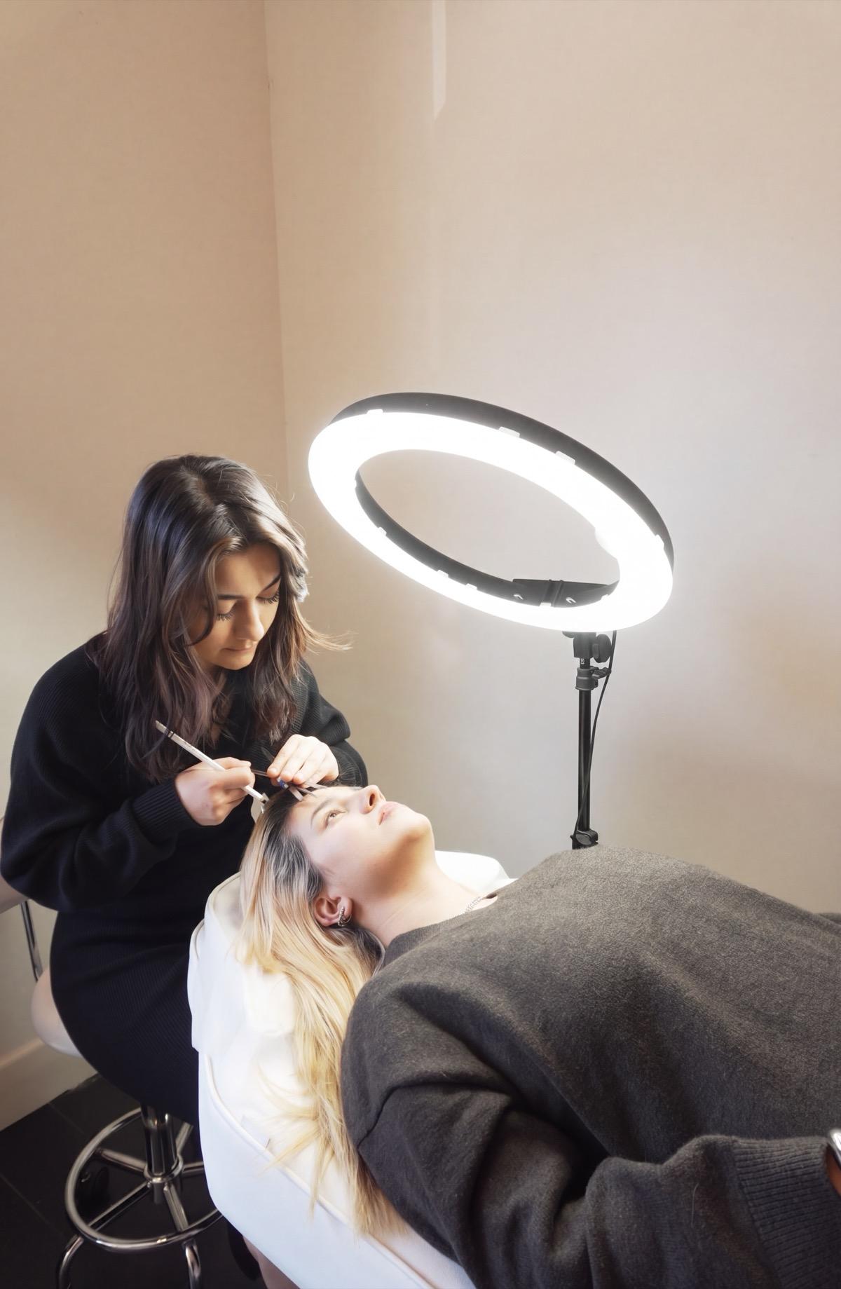 A brow artist shaping a guest's brows with a fine brush under a ring light in the upstairs studio