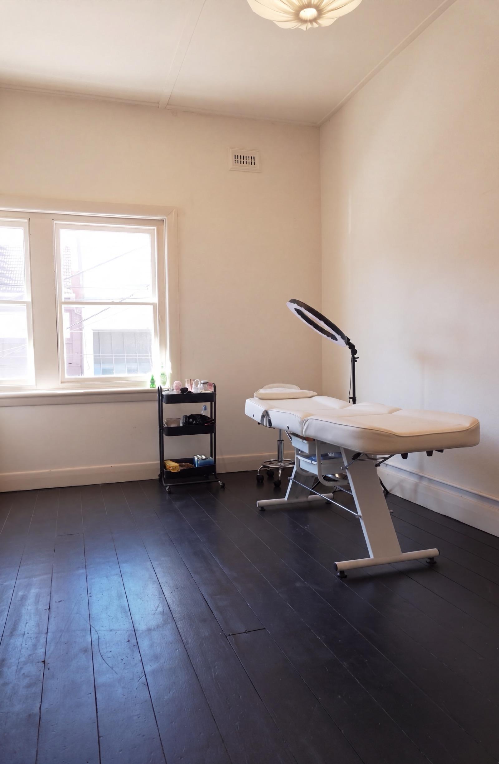 The upstairs brow studio: a white treatment bed and magnifying lamp beside a black trolley, window light across black floorboards